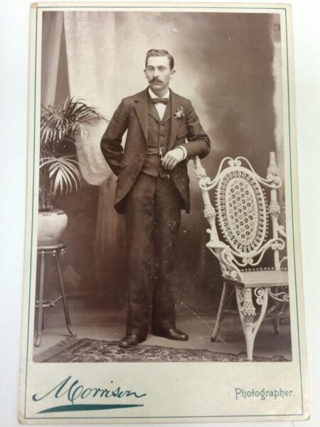 Photo of a man in a suit standing between a plant and a chair.