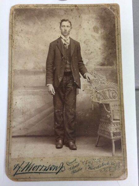 Old photo of a man in a suit standing next to a chair.
