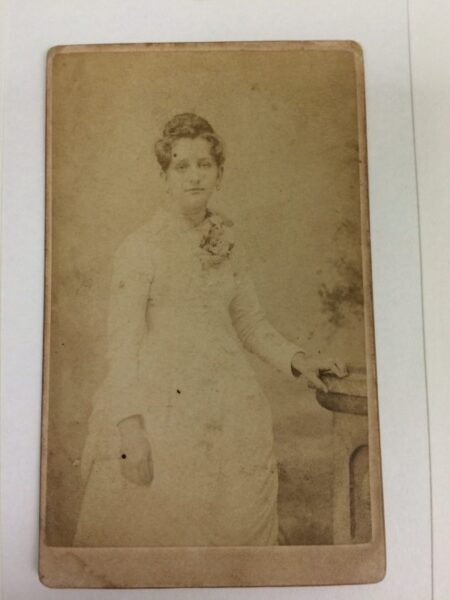 Old faded photo a woman in a white dress standing with hand on a table.