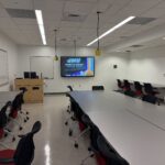 Image of classroom from the back of the room looking towards the front with a large row of tables and chairs.