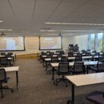 A view from the back of the classroom showing rows of tables and chairs and a two projector screen front.