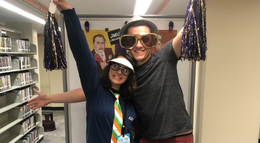 photo of two student employees with pom poms and costumes in front of library shelving