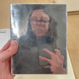 Hand holding a silver mirrored book. In the reflection, a blurry portrait of a person wearing glasses and holding the phone which captured the image.
