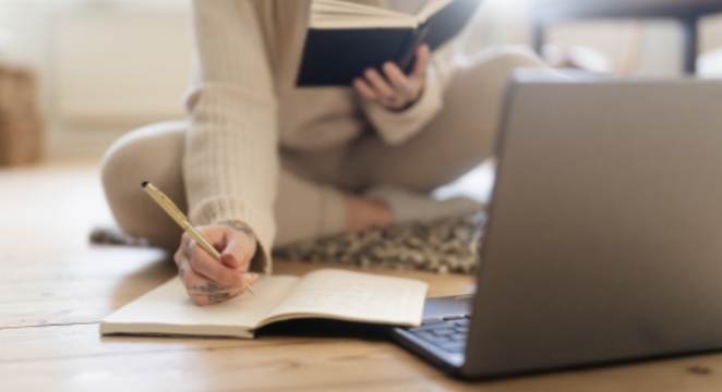 a person sits on the floor with an open book in hand. their other hand is writing in a notebook. an open laptop is in front of them.