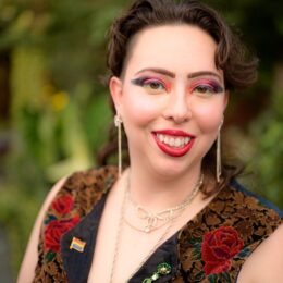 Photo shows Eva Pierce, a young white feminine person with short brown hair, orange makeup, a brown velvet vest with a rose pattern, and sparkly earrings and necklaces, and a frog pin on the right of their vest and a pride flag pin on the left of their vest. They are outside with a blurred background of green foliage.